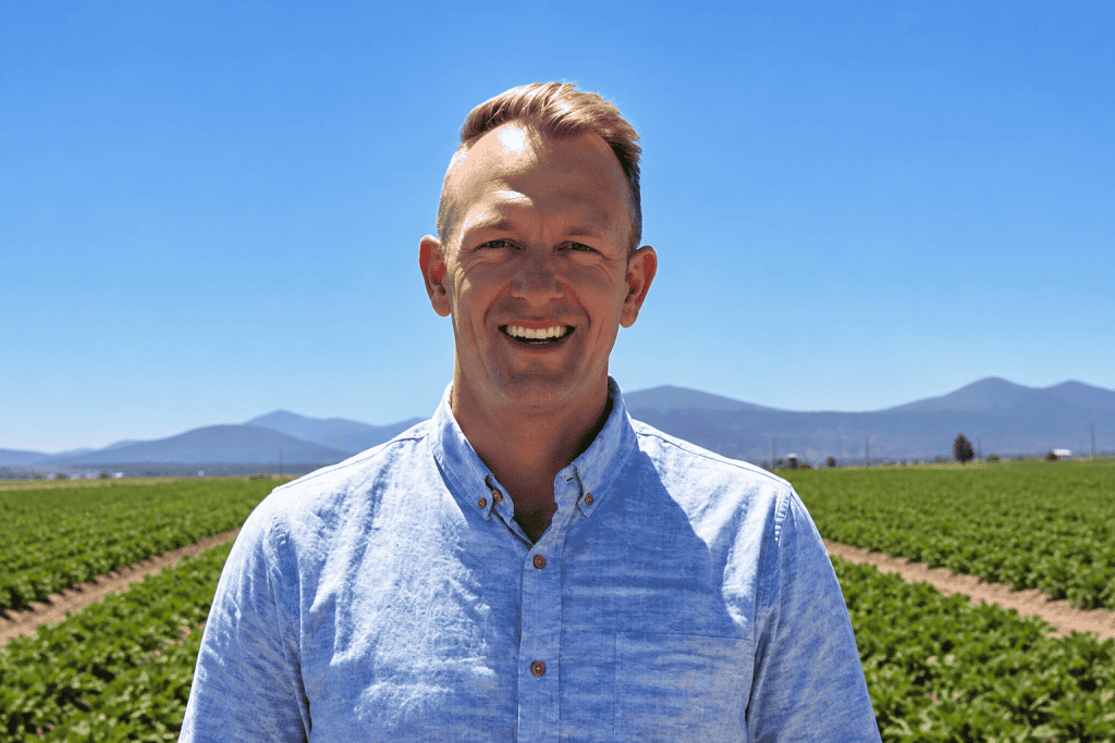 Smiling man in organic farm with green fields and mountains in background.