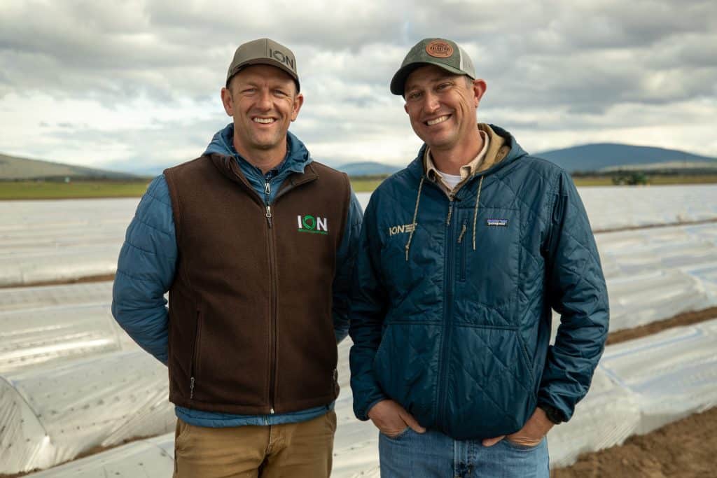 Two smiling men in outdoor farm setting with plastic-covered rows.