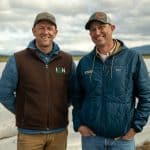 Two smiling men in outdoor farm setting with plastic-covered rows.